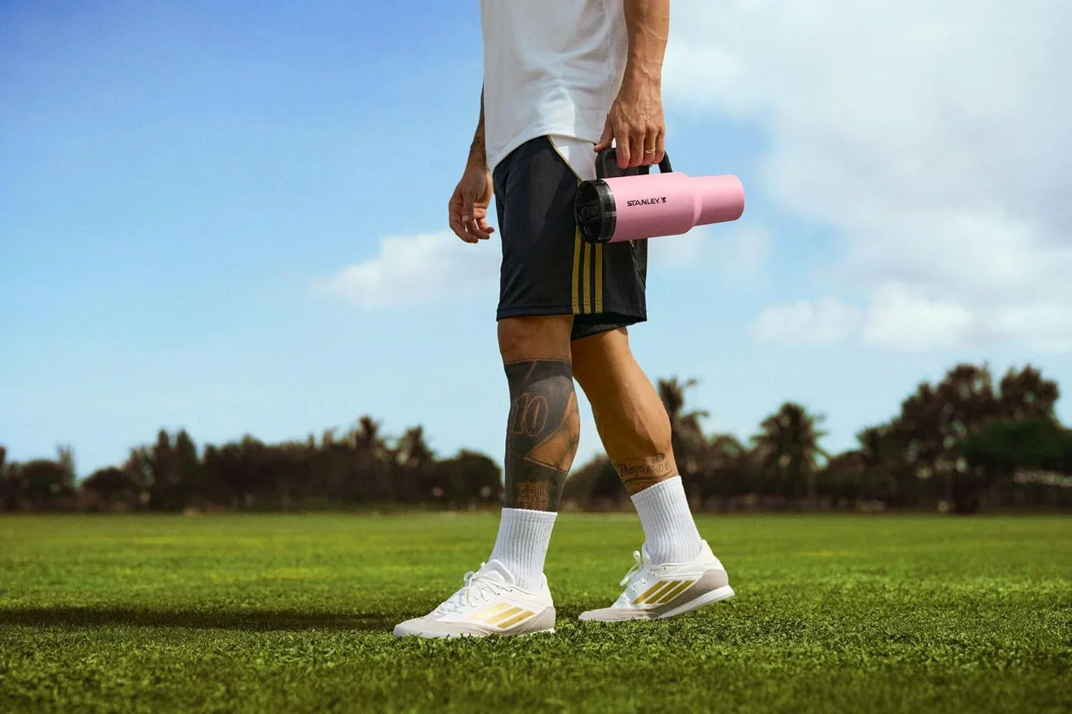 Person in sportswear holding a pink Stanley thermos on a green grass field under a blue sky