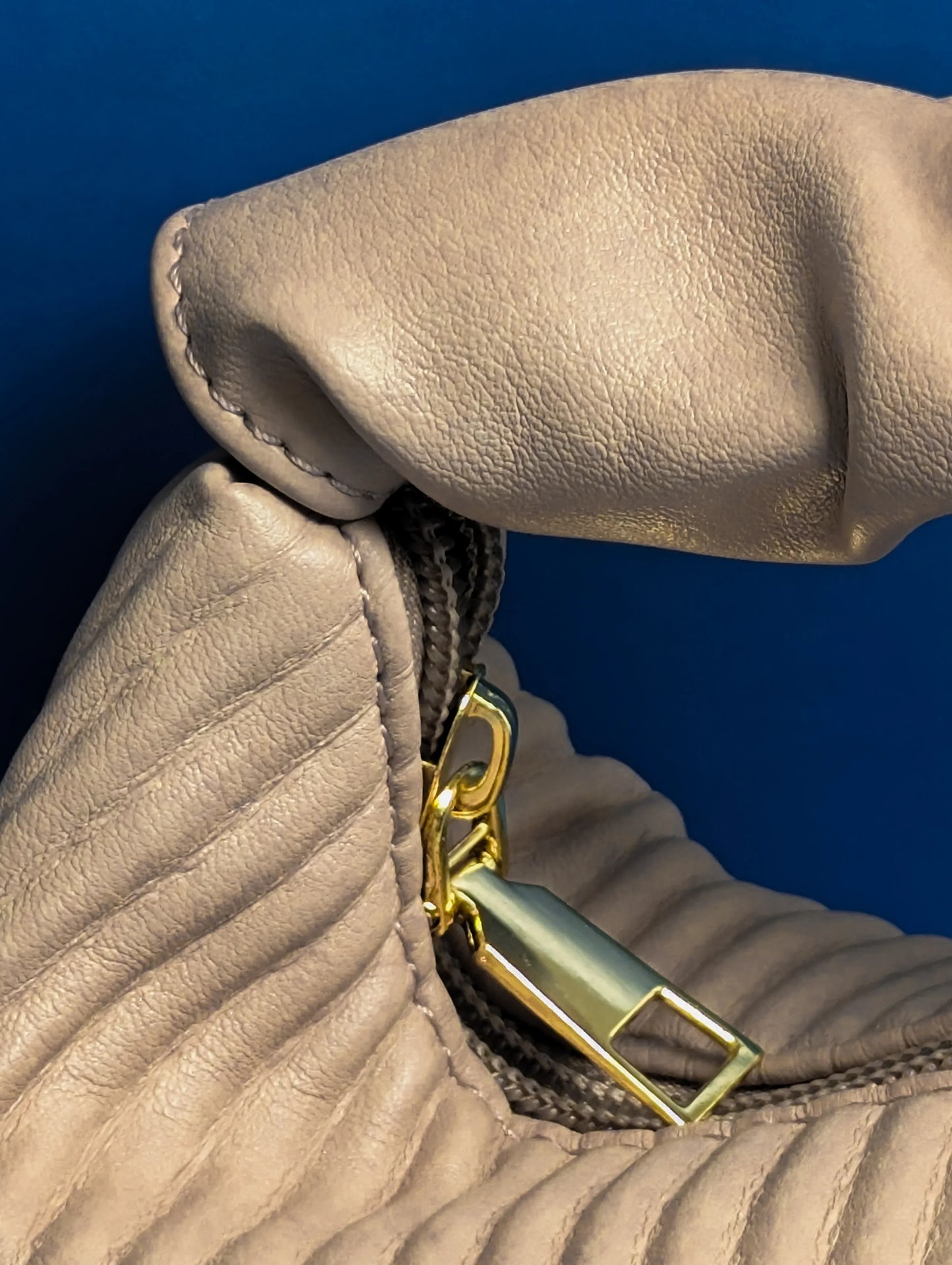 Beige leather handbag with gold zipper against blue background, showing detailed texture and stitching