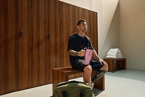 Man with tattoos sitting on wooden bench in locker room holding pink shaker bottle with green gym bag nearby