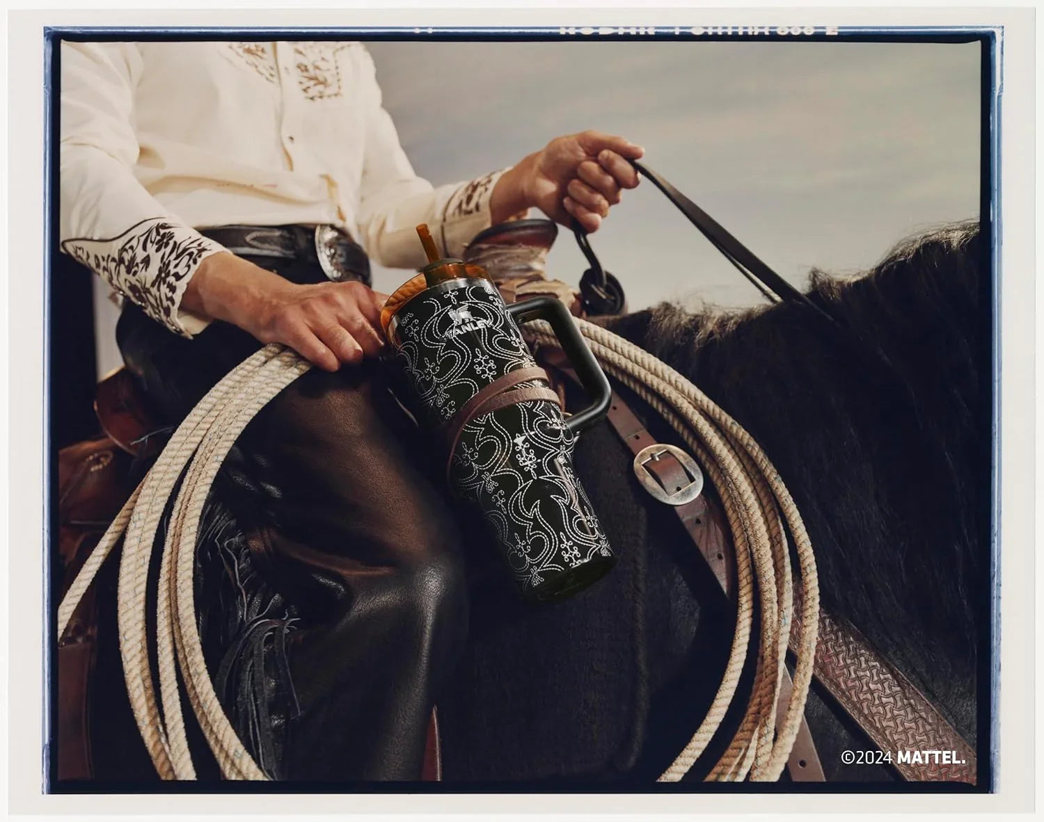 Cowboy in embroidered shirt and leather chaps holding reins on black horse with coiled rope and patterned thermos