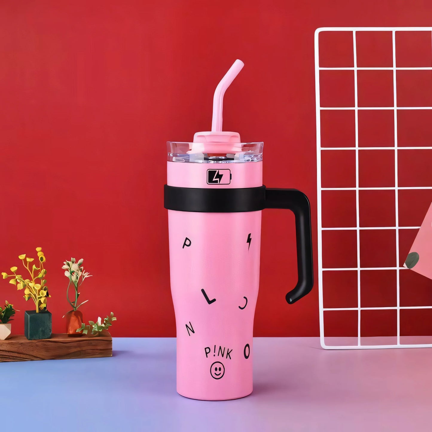 Pink tumbler with black handle and straw, decorated with scattered letters and smiley, on blue surface