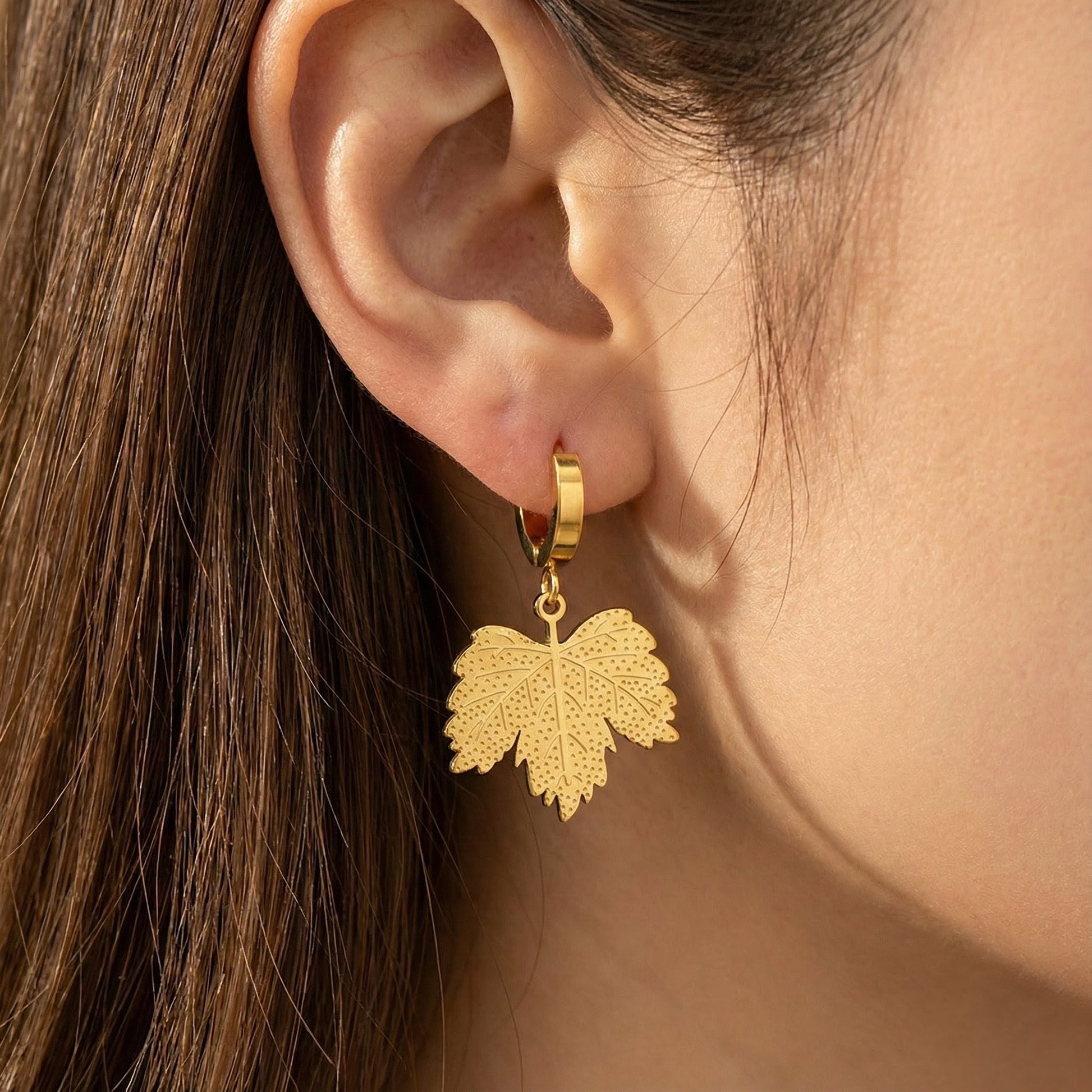 Close-up of golden leaf pendant hoop earring worn on ear, textured autumn jewelry