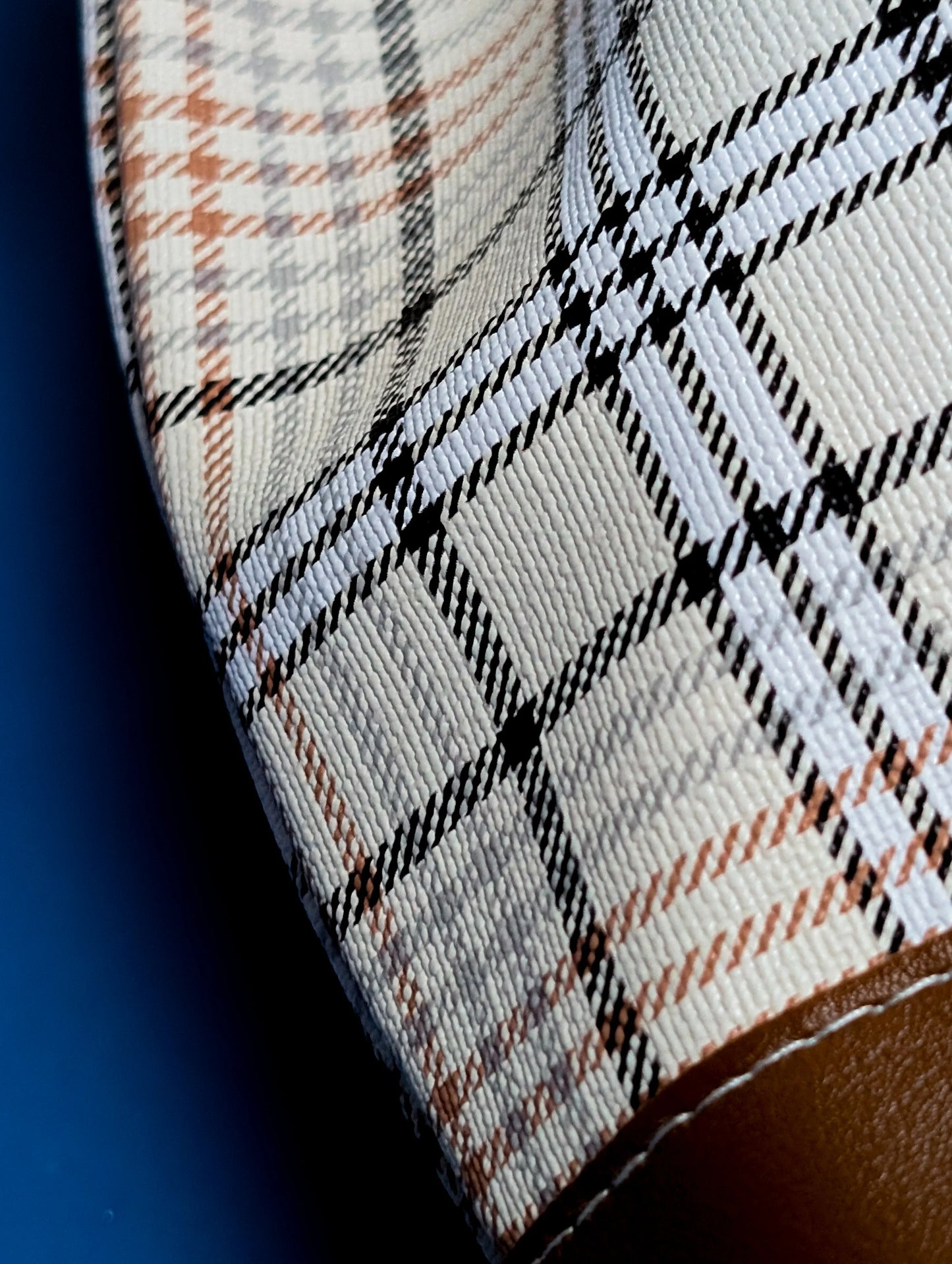 Close-up of beige checkered fabric with brown and black lines and brown leather trim on blue background