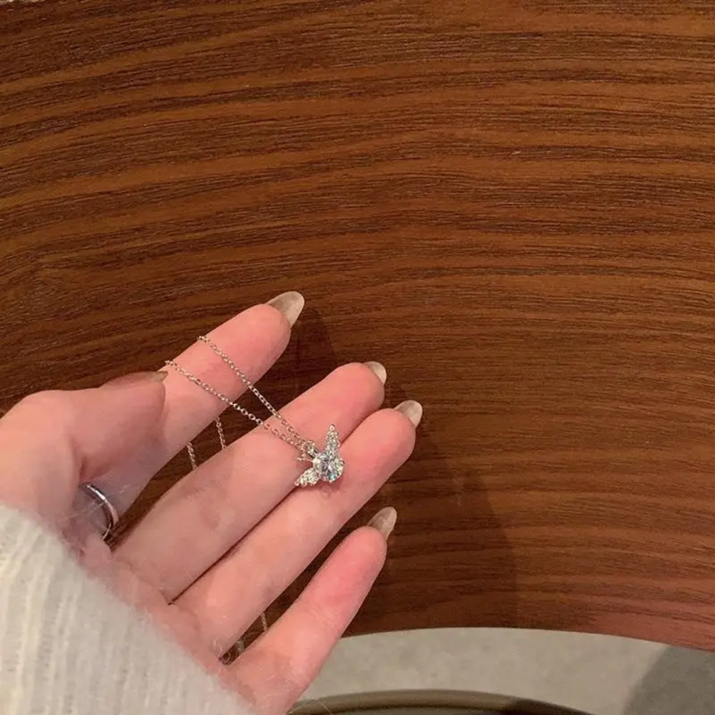 Hand holding silver chain necklace with butterfly-shaped crystal pendant over wooden surface