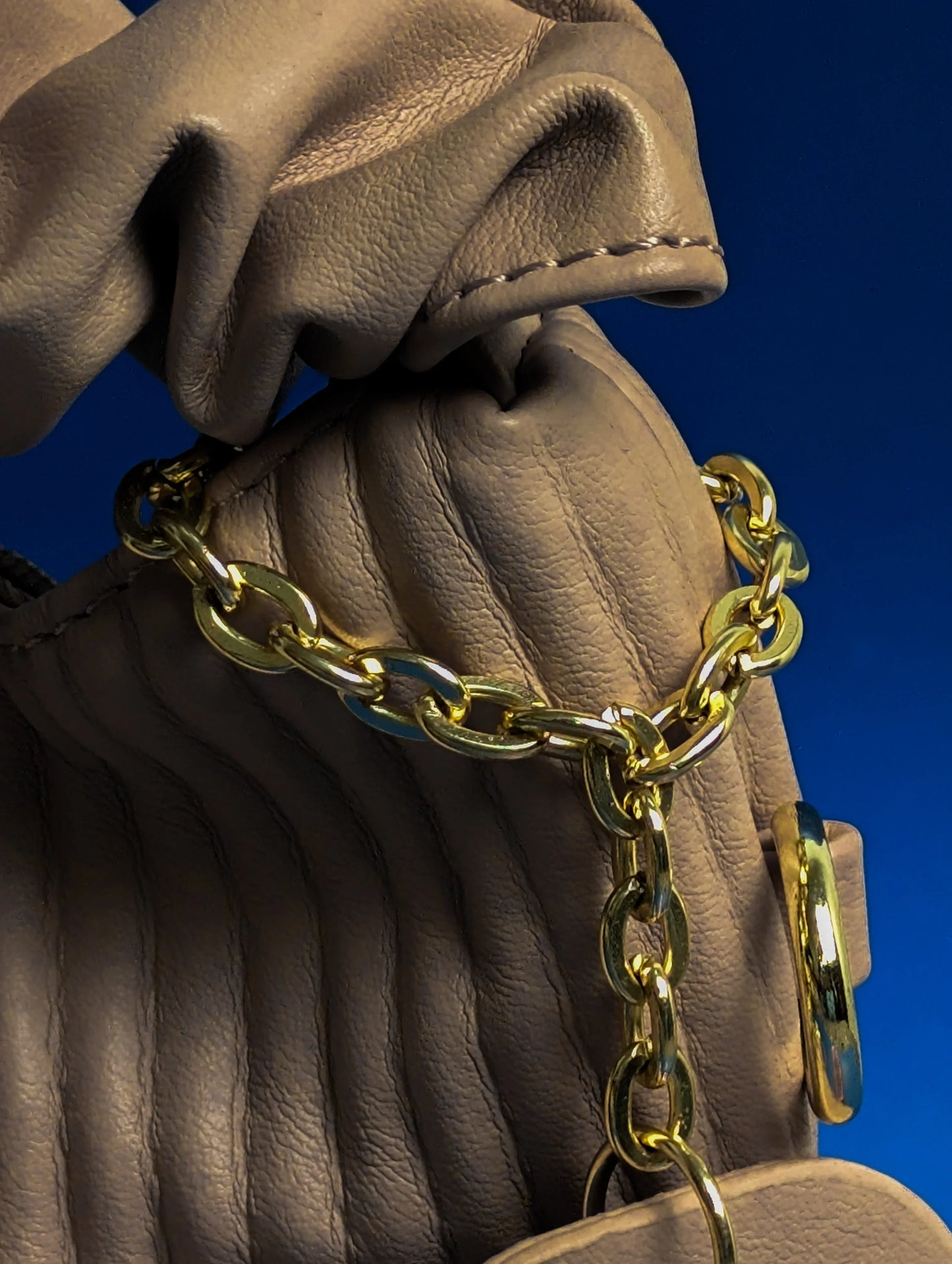 Close-up of beige leather handbag with ribbed texture and shiny gold chain strap on blue background
