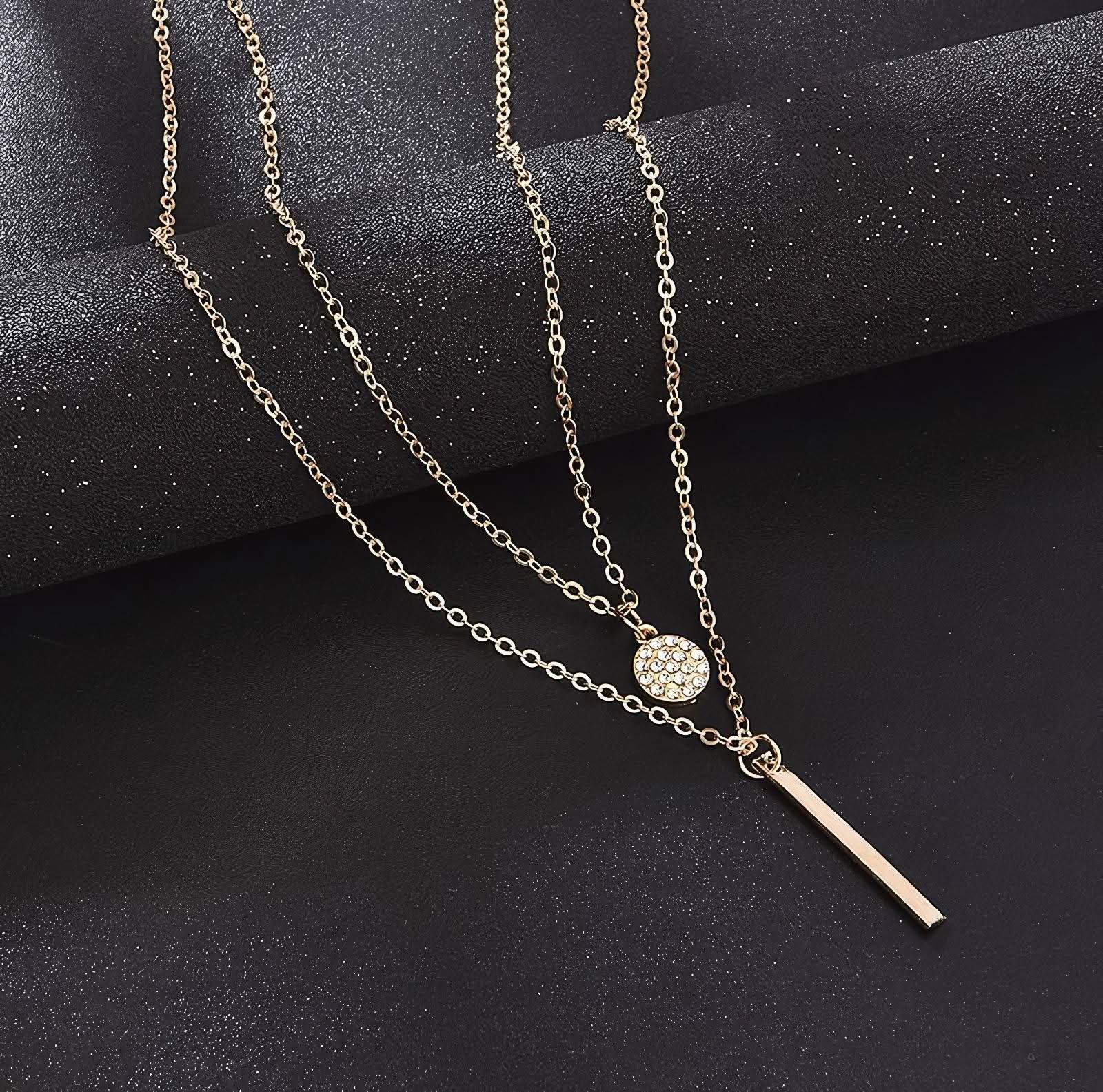 Double-layer gold chain necklace with round rhinestone pendant and vertical bar pendant on black textured background