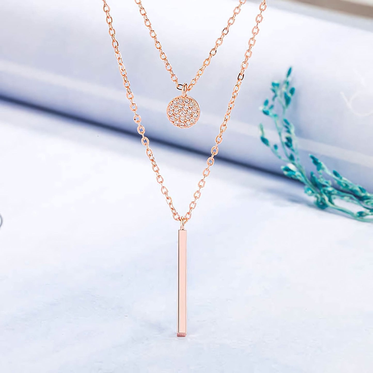 Two-layer rose gold necklace with round pendant and vertical bar on light surface with green plant
