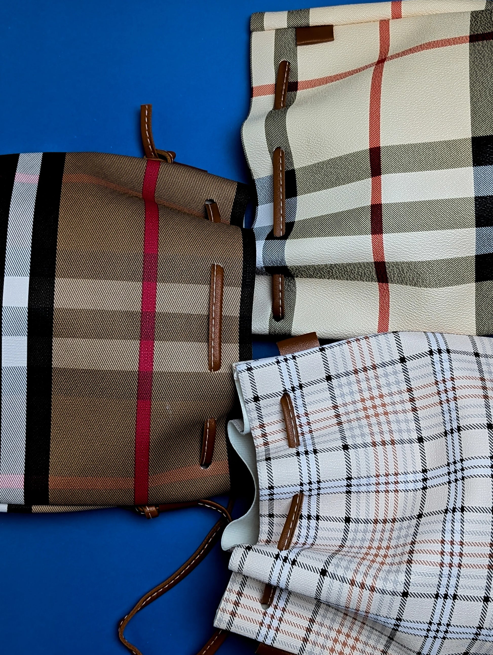 Three plaid drawstring bags with leather details on a blue background in beige, brown, and white tones