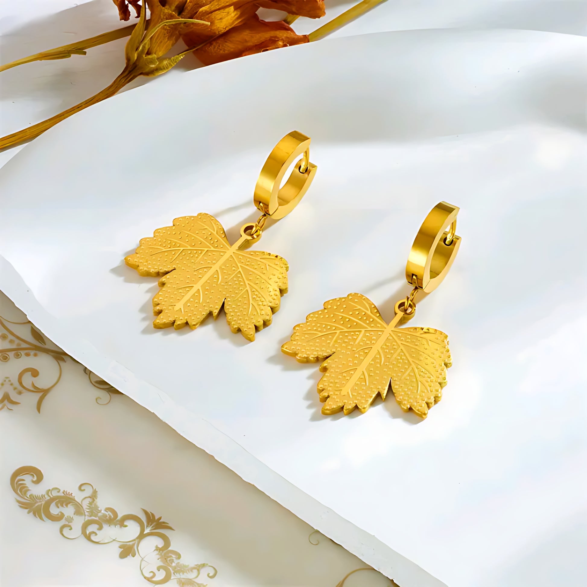 Gold leaf-shaped dangle earrings on white surface with decorative dried orange flowers