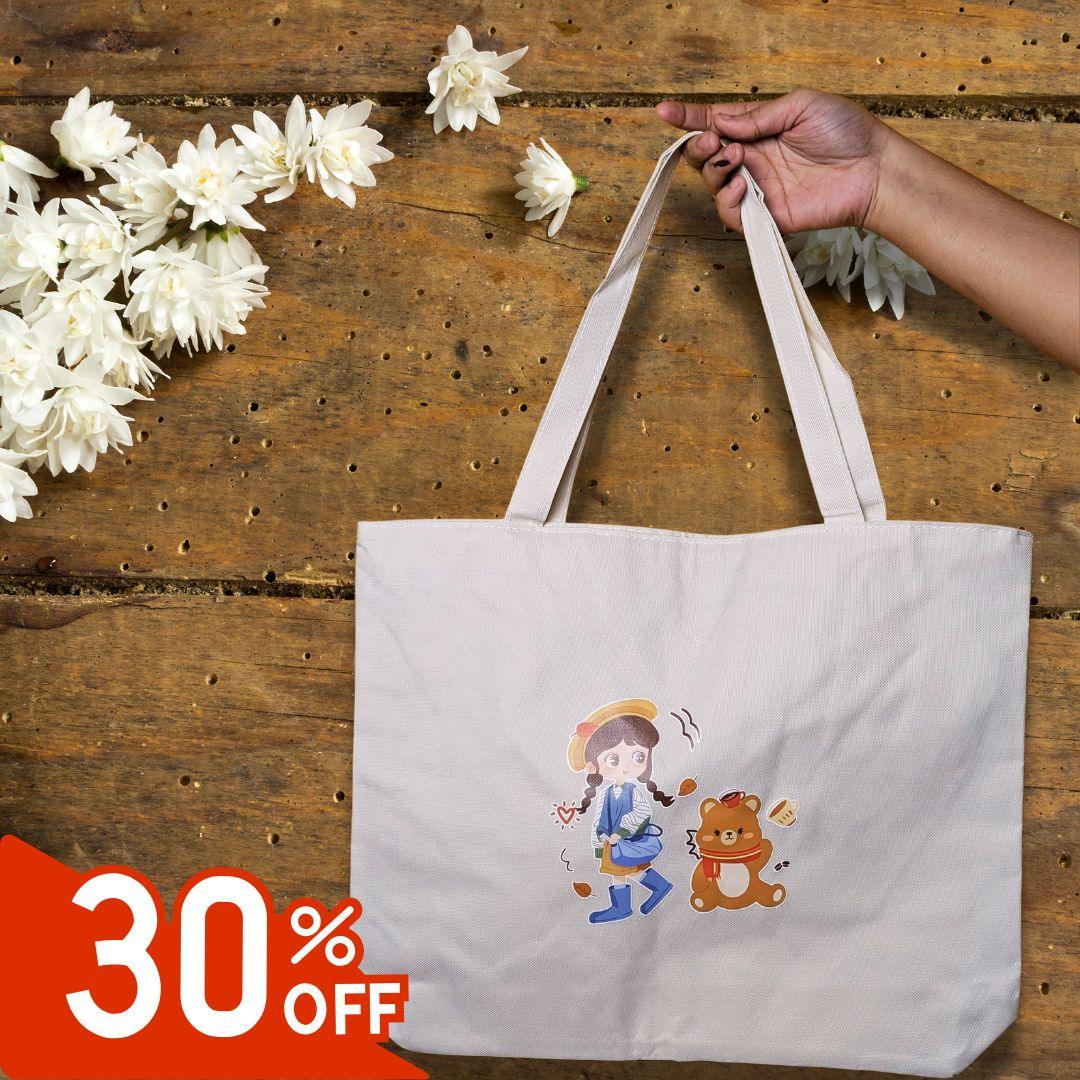 Hand holding beige tote bag with cartoon girl and bear design on wooden table with white flowers and 30% off label