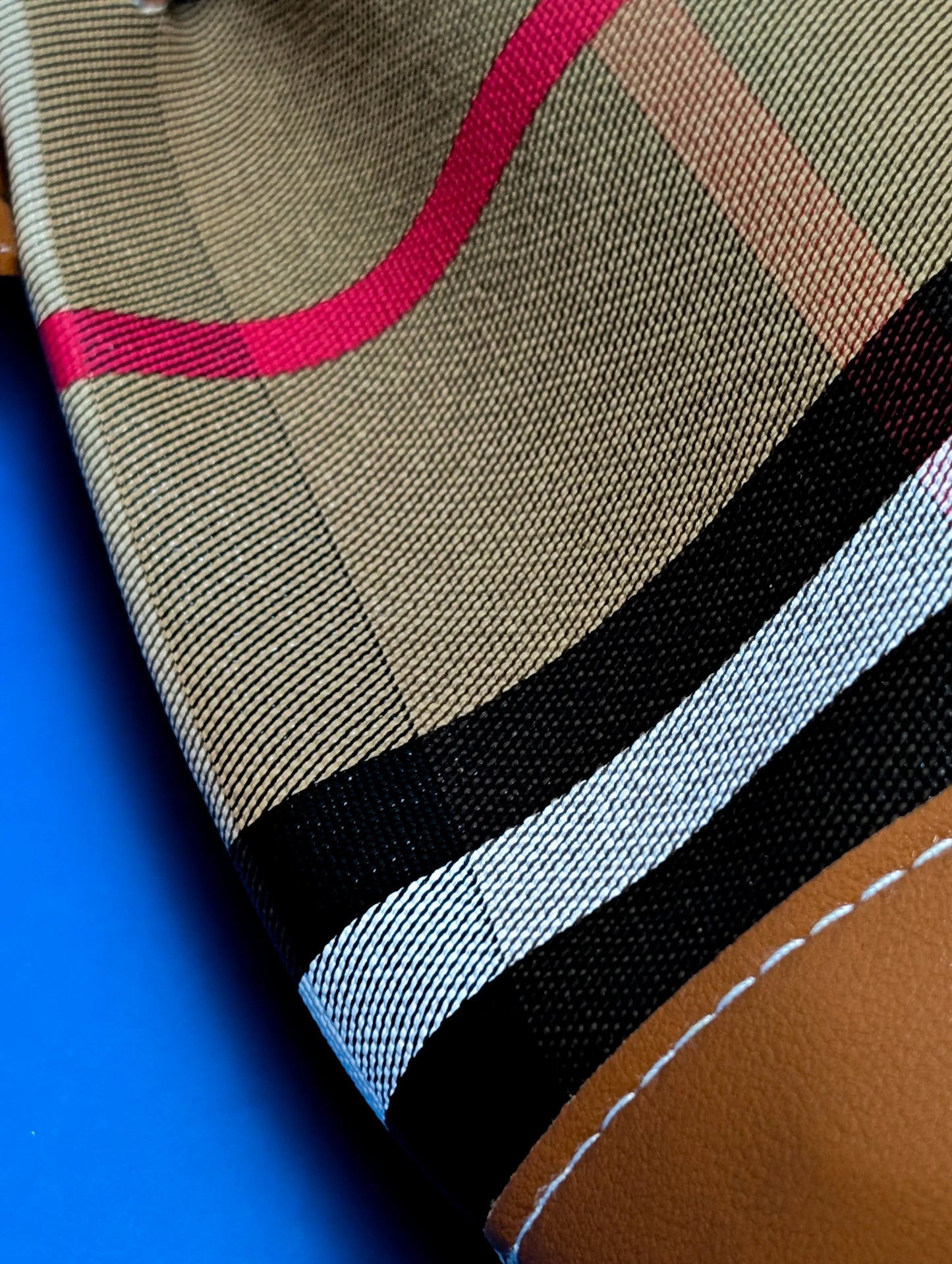 Close-up of beige plaid fabric with pink, black, white stripes and brown leather patch on blue background