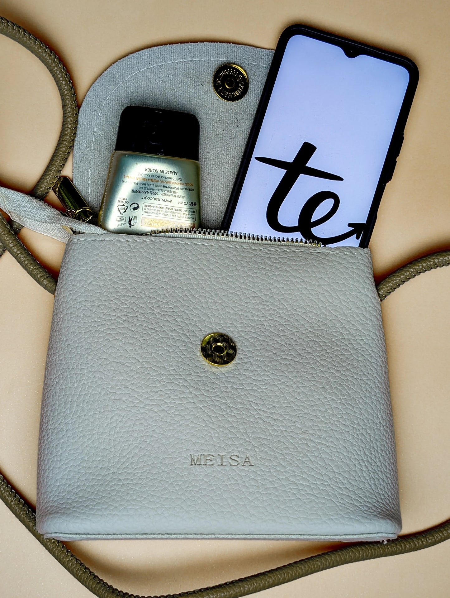 Gray MEISA leather crossbody bag with a smartphone and hand sanitizer inside on beige surface