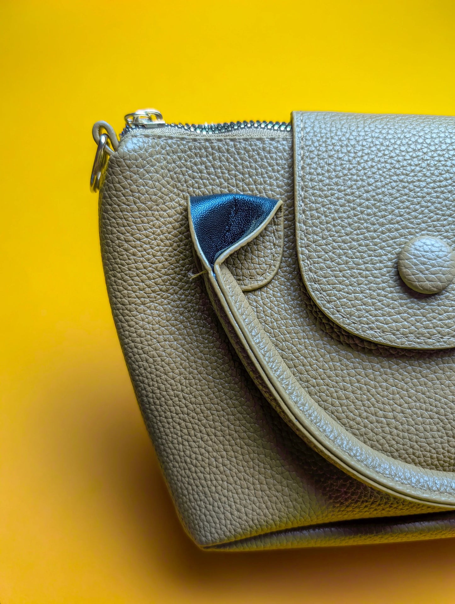 Close-up of a textured taupe leather handbag with zipper and button flap on a bright yellow background