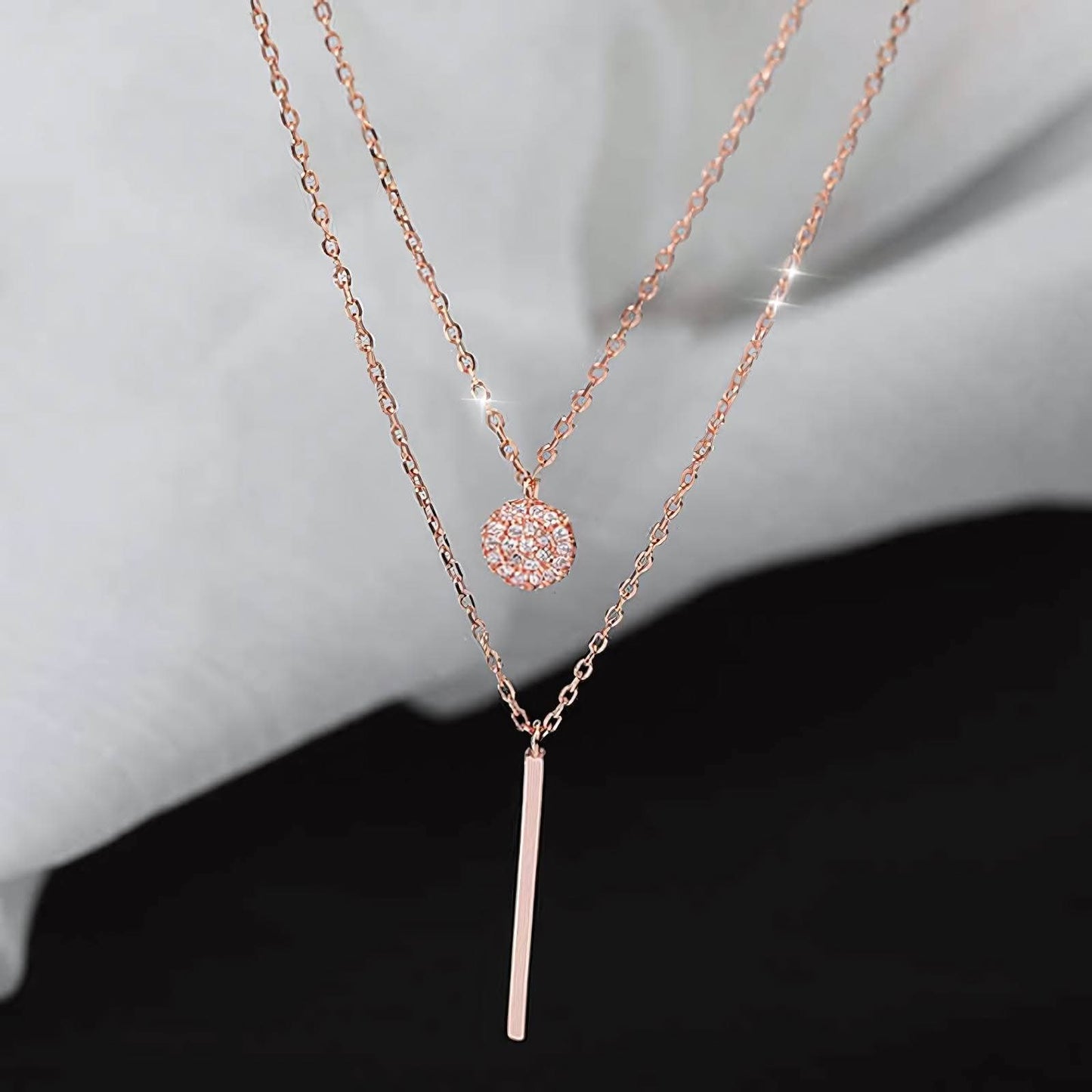 Elegant layered rose gold necklaces with a sparkly round pendant and a sleek bar pendant on a soft gray background