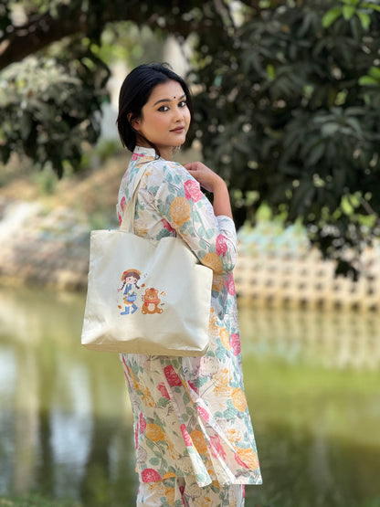 Woman in floral kurta holding beige tote bag with cartoon girl and bear print by water