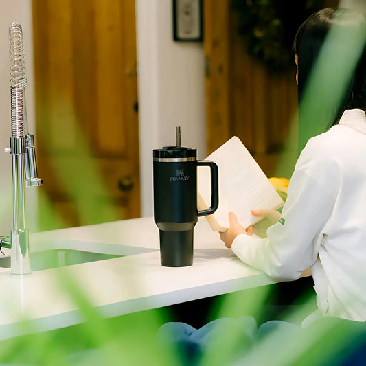 Black Stanley insulated tumbler on white kitchen counter near woman reading a book by wooden door