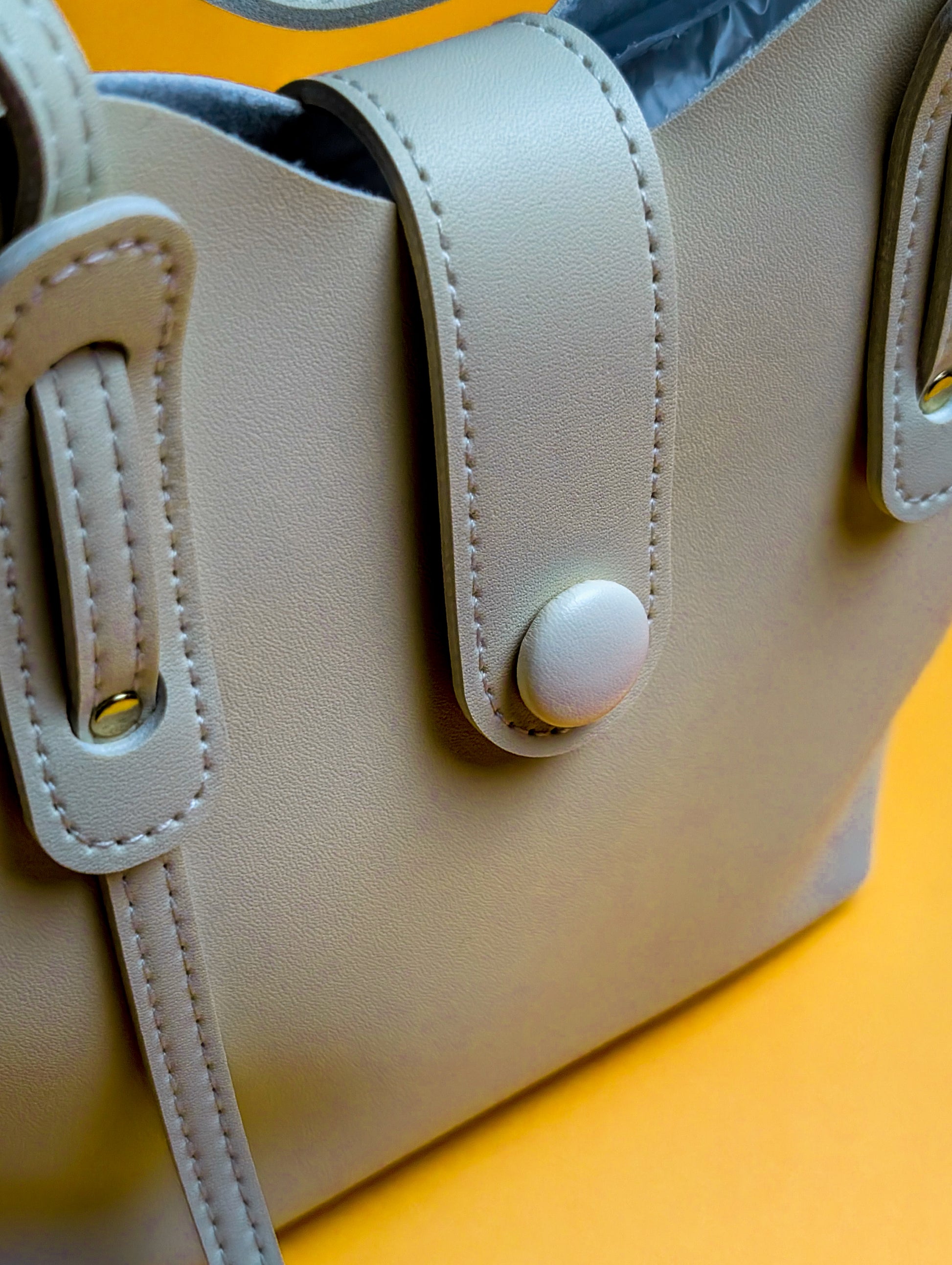 Close-up of beige leather handbag with stitched straps and button detail on yellow background