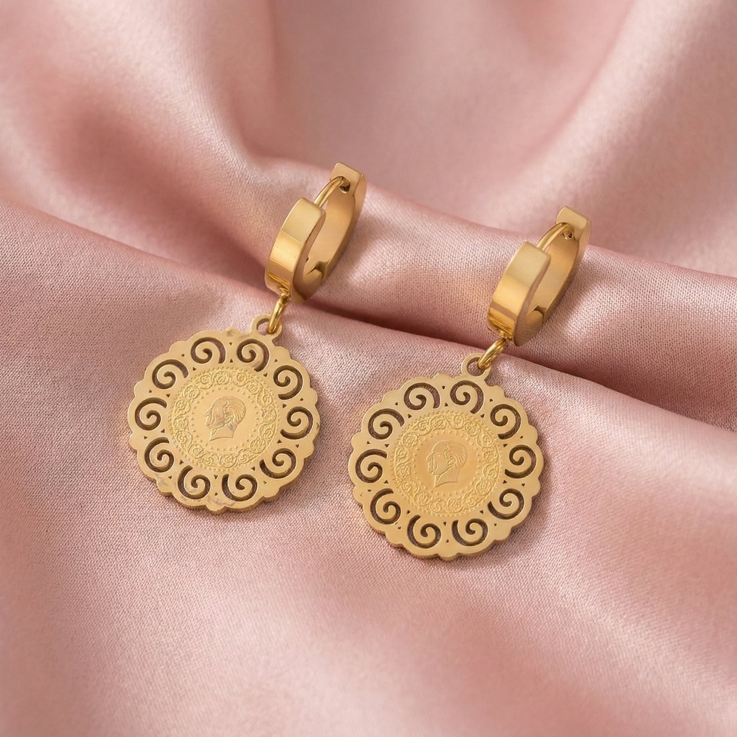 pair of gold hoop earrings with intricate coin-shaped pendants on pink satin fabric