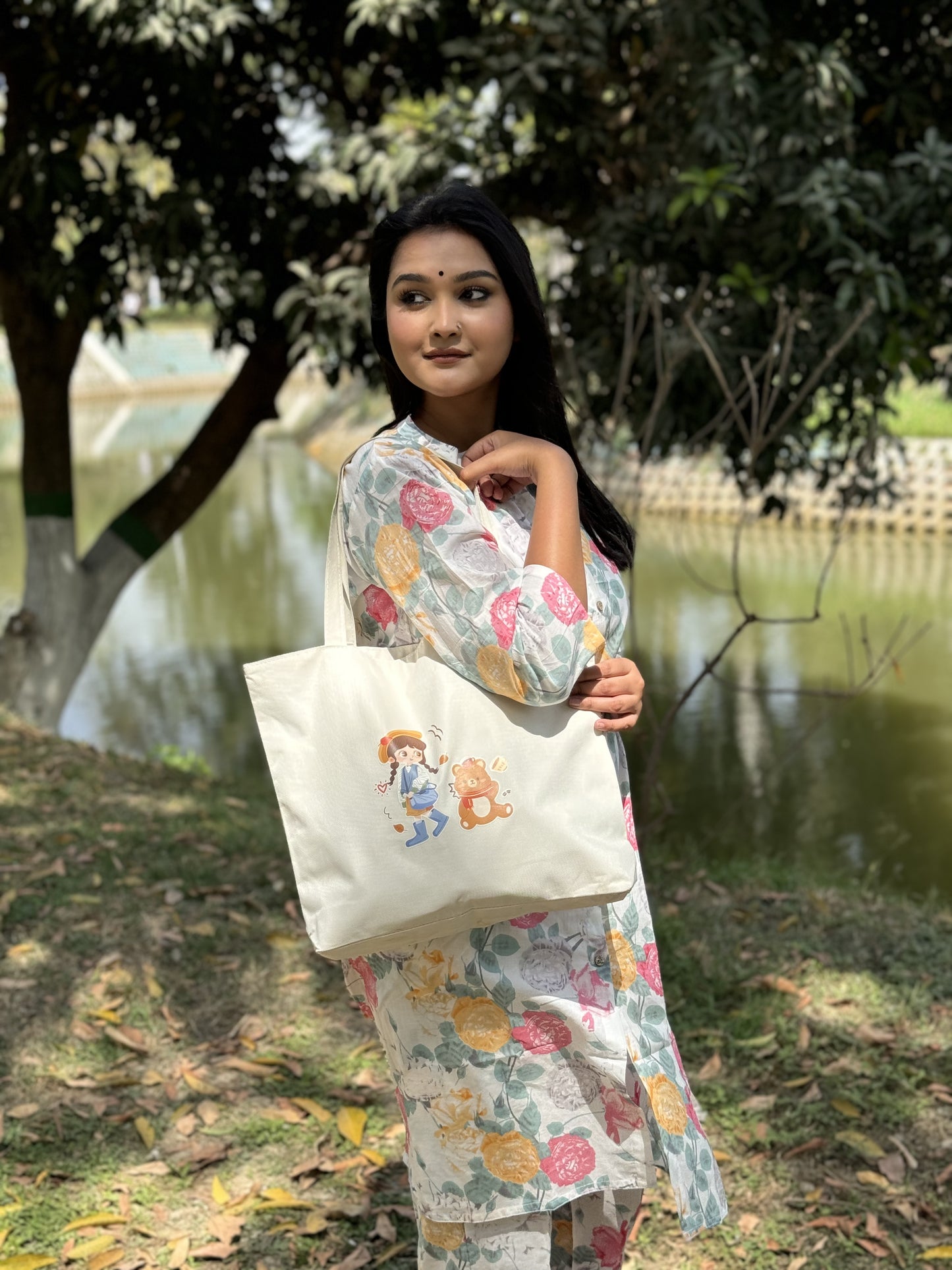Woman in floral kurta holding white tote bag with cartoon girl and bear by lakeside