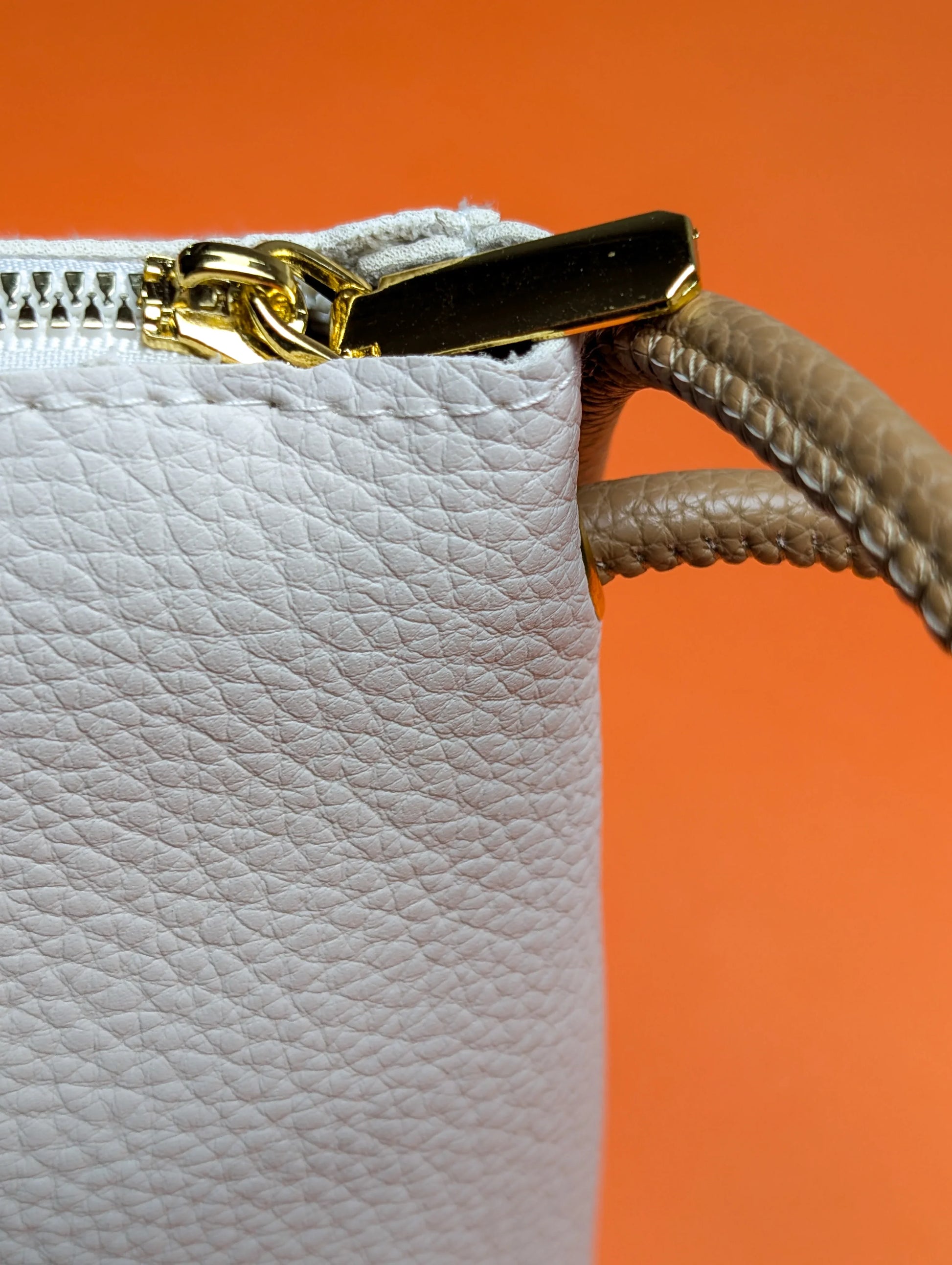 Close-up of white textured leather handbag with gold zipper and beige braided handle on orange background