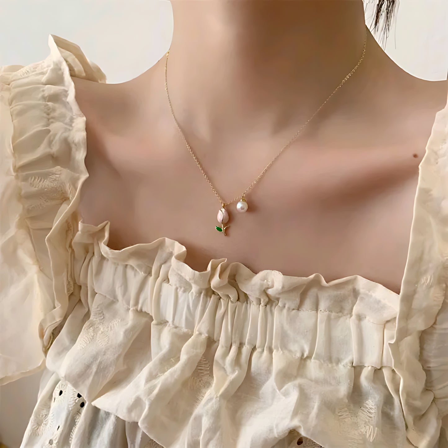 Close-up of woman wearing gold necklace with pink flower and pearl pendant, in a cream ruffled top