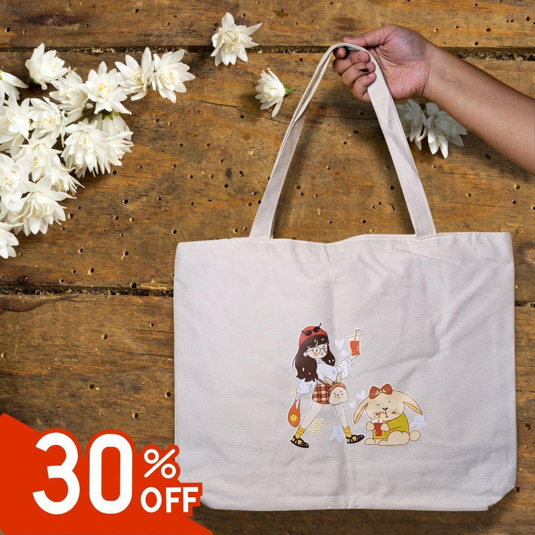 Hand holding beige tote bag with cartoon girl and bunny design on rustic wooden table with white flowers and 30% off tag